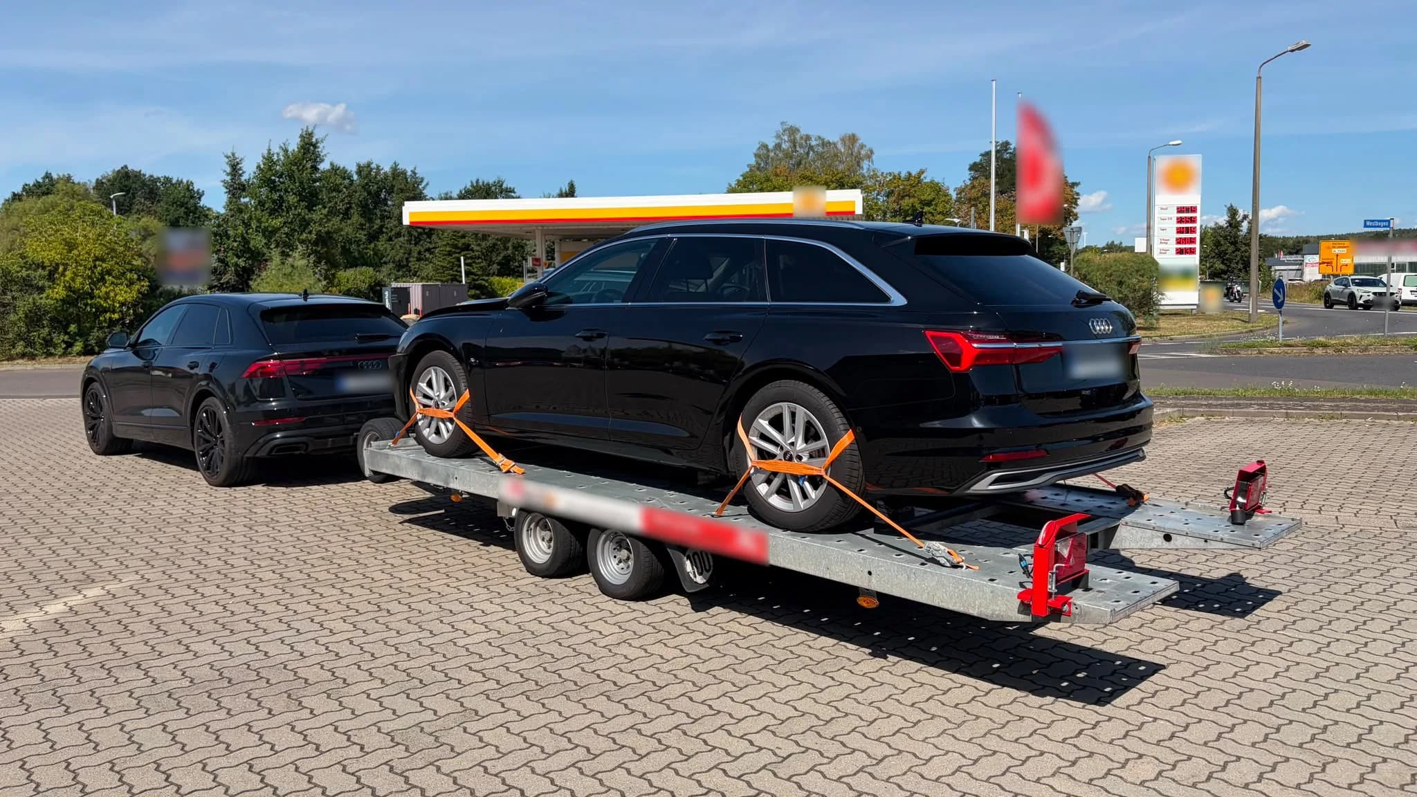 Schwarzer Audi A6 Avant, mit Spanngurten auf einem zweiachsigen Autoanhänger gesichert, gezogen von einem Audi Q8